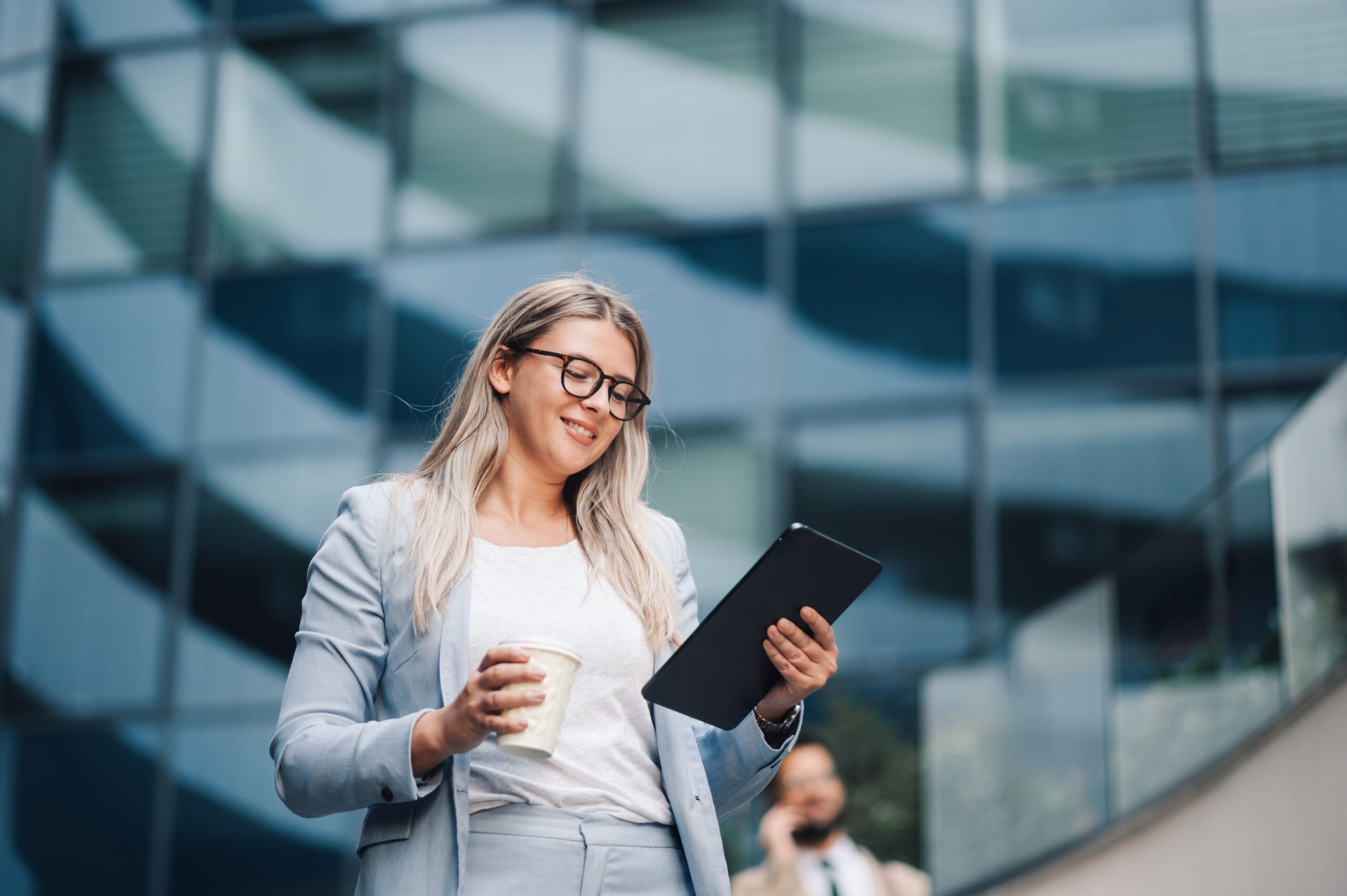 Businesswoman is holding a coffee cup and using a digital tablet with a smile on her face while standing outside of an office building