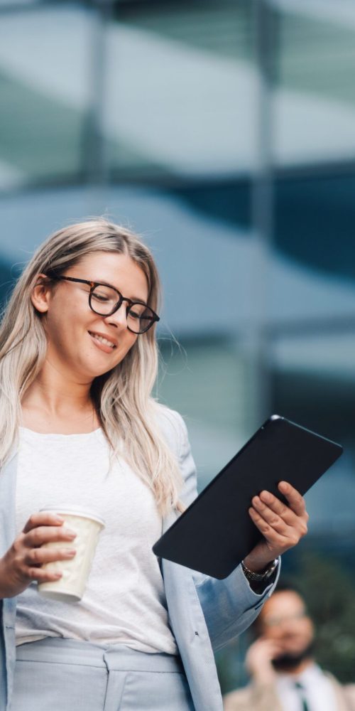 Businesswoman is holding a coffee cup and using a digital tablet with a smile on her face while standing outside of an office building
